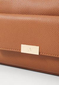 Sac en cuir texturé marron avec une plaque de matériel doré lisse présentant un logo en relief. Lignes horizontales distinctives sur la surface.