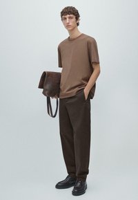 Camicia marrone a maniche corte abbinata a pantaloni larghi marrone scuro. Nella mano è tenuta una borsa marrone testurizzata. Le calzature sono scure e eleganti.