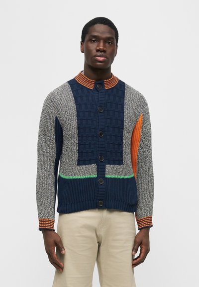 Άνδρας που φοράει πολύχρωμη πλεκτή ζακέτα με κουμπιά στο μπροστινό μέρος, σε συνδυασμό με ανοιχτό μπεζ παντελόνι, στέκεται μπροστά σε ένα απλό φόντο.