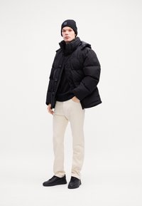 Puffer jacket in black, with a high collar and snap buttons. Worn over a black sweater, paired with cream-colored pants and black sneakers.