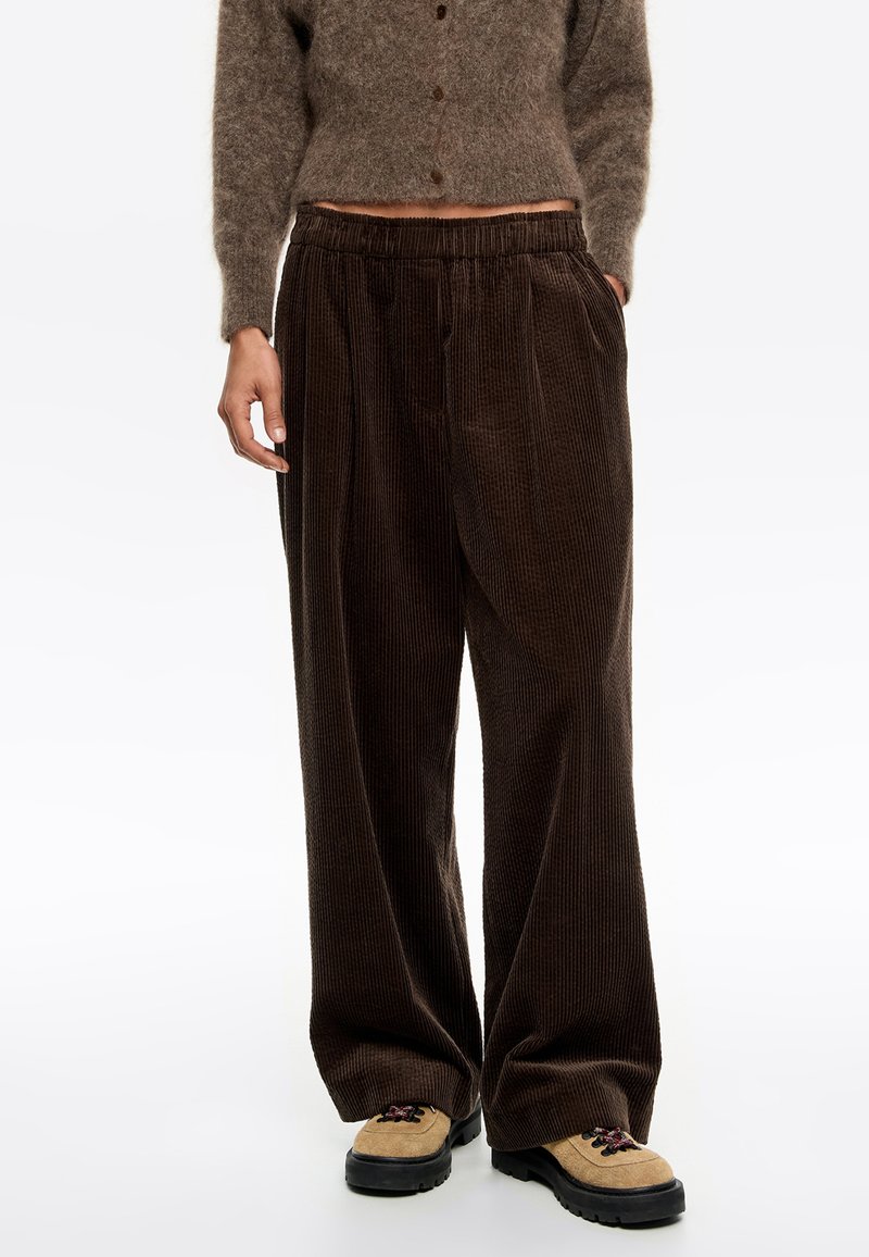 Persona que lleva pantalones de pana marrón oscuro holgados, zapatos de senderismo beige y un cárdigan corto de lana marrón con botones.