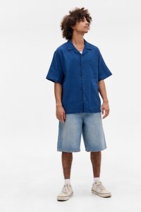 Camicia azzurra a maniche corte con bottoni e taschino, abbinata a pantaloni corti di jeans azzurri chiari. Il modello indossa sneakers bianche con calzettoni chiari.