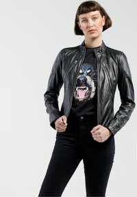 Schwarze Lederjacke mit tailliertem Design und Reißverschluss. Getragen über einem schwarzen T-Shirt mit grafischem Bild eines Hundegesichts. Schwarze hochgeschnittene Hose.