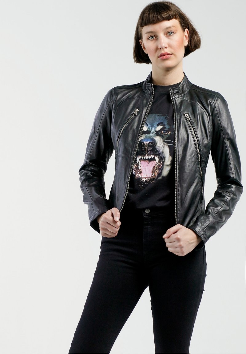 Schwarze Lederjacke mit tailliertem Design und Reißverschluss. Getragen über einem schwarzen T-Shirt mit grafischem Bild eines Hundegesichts. Schwarze hochgeschnittene Hose.