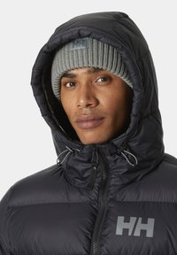 Zwarte pufferjack met een hoge kraag, grijze gebreide beanie met een logo-label en trekkoorddetails op de capuchon. Glad textuur, modern ontwerp.