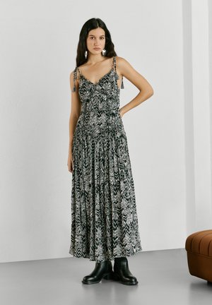 Mujer con cabello largo y oscuro, vestida con un vestido maxi sin mangas con estampado en blanco y negro, y botas negras hasta el tobillo, de pie con una mano en la cadera.