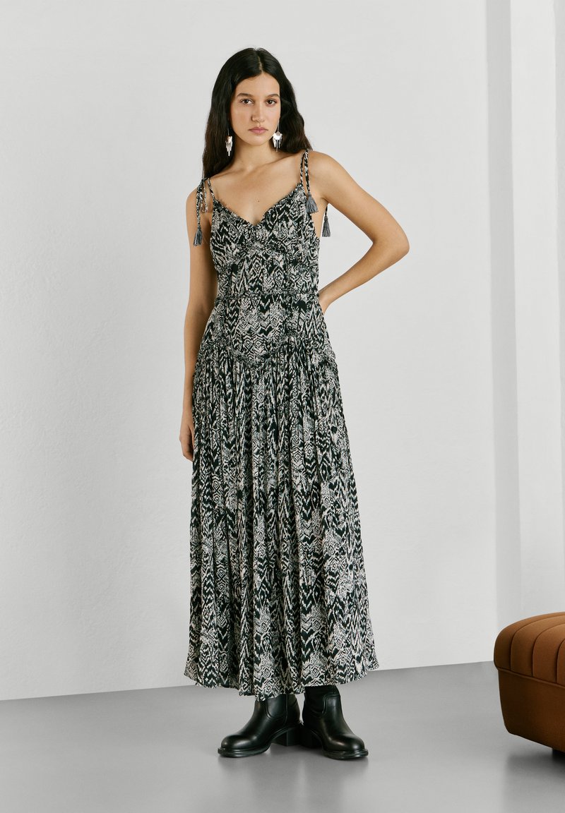 Moteris su ilgais tamsiais plaukais, vilkinti juodai baltą, be rankovių maximalaus ilgio suknelę su raštu, juodais aulinukais, stovinti viena ranka ant klubų.