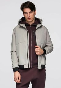 Młody mężczyzna w jasnoszarej kurtce z kapturem, noszący ciemnoczerwony polar na zamek i pasujące spodnie, stojący na tle jednolitego koloru.