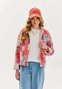 Prešívaná patchworková bunda v červenej, ružovej a kvetinových vzoroch, so zipsom vpredu a dvoma bočnými vreckami. Skombinovaná s ľahkým tričkom a džínovými nohavicami.