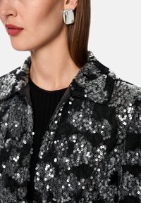 Black sequined jacket with a collar and intricate lace detailing. Model wears a silver square earring. Background is plain white.