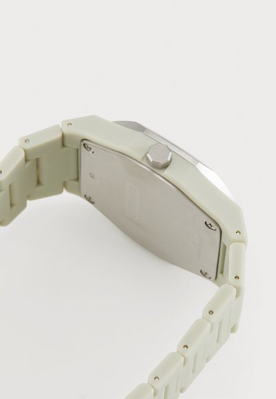 Montre carrée avec un bracelet en silicone vert pâle mat et un dos en acier inoxydable. Présente une couronne hexagonale en argent et une texture de surface lisse.