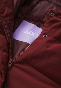 Burgundská bunda má zipovou kapsu s fialovým štítkem s nápisem "JJX." Tkanina má jemnou mřížkovou texturu a patentky.