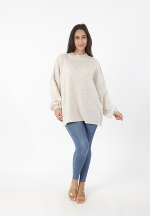 Suéter de punto beige de gran tamaño con cuello alto y hombros caídos, combinado con jeans ajustados azules y sandalias transparentes con tiras.
