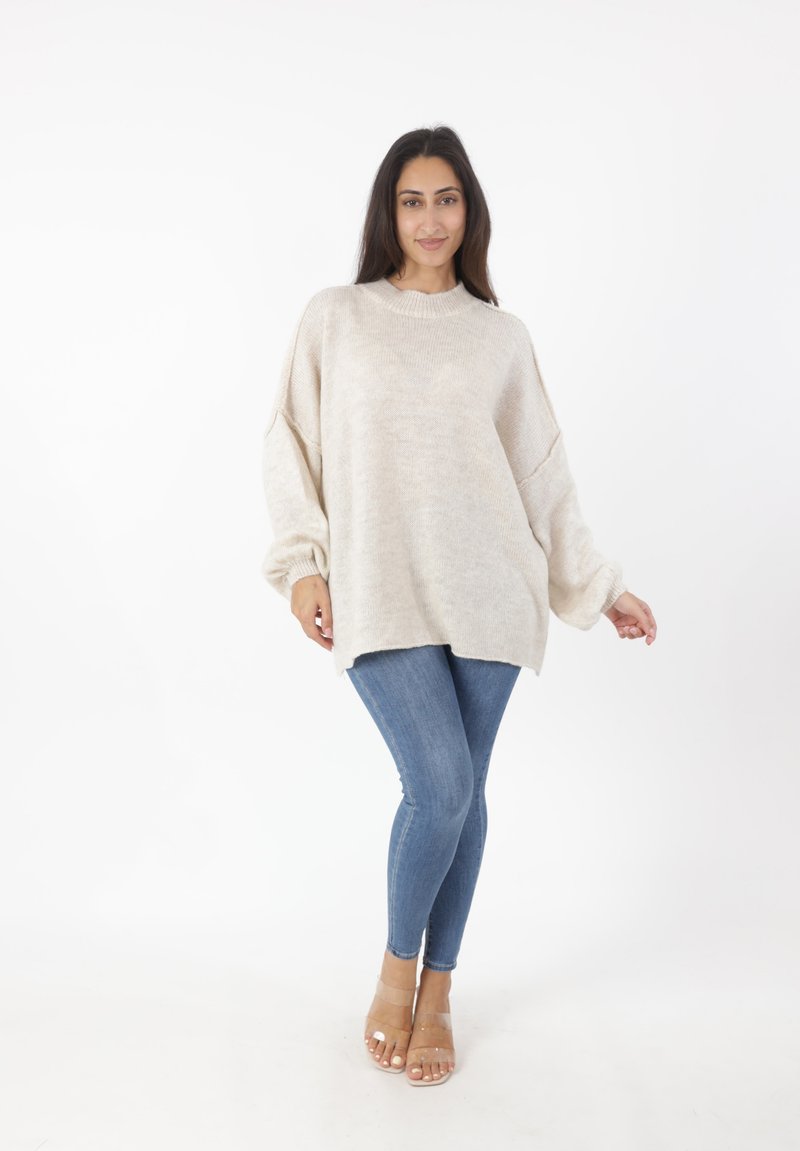 Maglione oversize beige a collo alto con spalle scese, abbinato a jeans skinny blu e sandali trasparenti con cinturini.