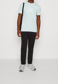 Camiseta de algodón azul claro con logo blanco, pantalones negros y zapatillas grises con detalles negros; pose de pie contra un fondo neutro.