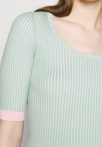 Haut en maille côtelée vert clair avec un large décolleté rond et des manches courtes ornées d'accents roses aux poignets. Texture lisse.