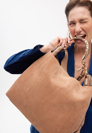 Donna con giacca blu tiene e morde una grande borsa beige a coste con manico metallico rotondo su sfondo bianco.