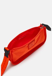 Pomarańczowa nylonowa torba z zamkiem błyskawicznym, o gładkiej fakturze i małej naszywce z logo. Wnętrze wyłożone jednolitą czerwoną tkaniną.