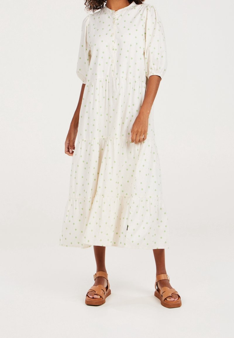 Femme portant une robe midi blanche à imprimé floral vert et manches bouffantes, associée à des sandales marron à bride chevillère, debout sur un fond blanc.
