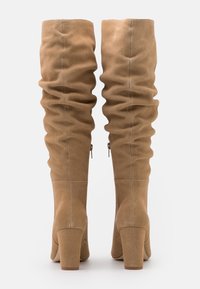 Bottes marron en daim montant jusqu'aux genoux avec une texture plissée, un talon bloc et un détail de fermeture éclair latérale. Surface lisse avec des accents de coutures subtiles.