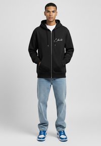 Haut à capuche zippé noir avec cordons, poches avant et logo en script blanc. Associé à un jean bleu clair et des baskets bleues.