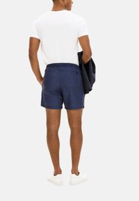 Shorts di lino blu navy con vita elasticizzata, dettaglio della tasca posteriore, indossati con una t-shirt bianca e scarpe bianche. Design minimalista.