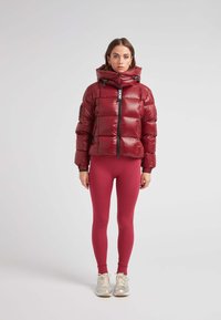 Giacca a vento rossa con finitura lucida, collo alto e chiusura con zip; indossata sopra leggings aderenti a coste color bordeaux e sneakers beige.