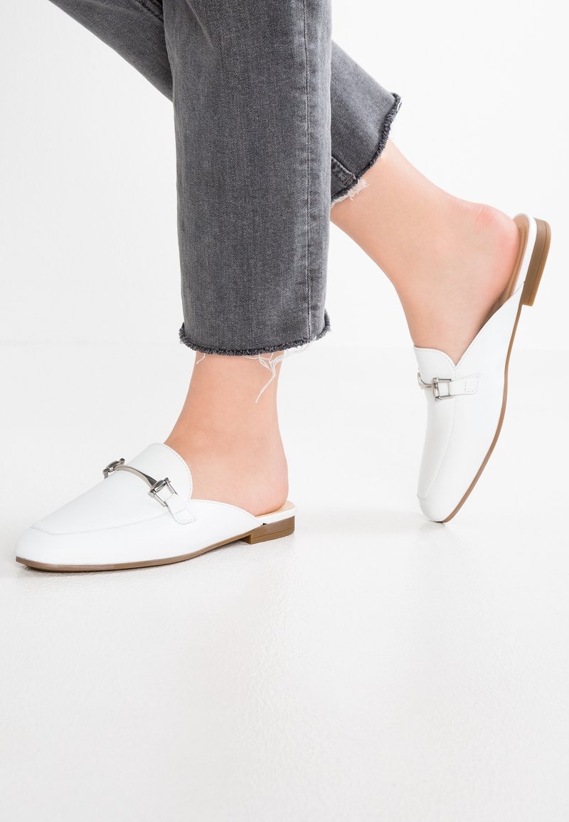 Personne portant des mocassins blancs sans lacets avec un détail de boucle en métal et un jean gris à ourlet effiloché sur un sol blanc.