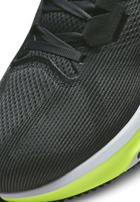 Zapatilla deportiva negra con una parte superior de malla texturizada, con acentos grises y una suela verde vibrante. Los cordones son negros, con un diseño aerodinámico.