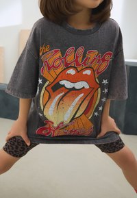 Szürke túlméretezett póló, amelyen a Rolling Stones grafikája látható, piros nyelv logóval és narancssárga részletekkel, fekete leopárdmintás leggingsszel párosítva.