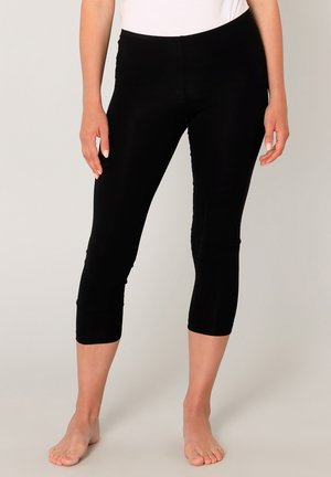 Vrouw draagt zwarte korte legging en een wit topje, staat op blote voeten tegen een effen lichte achtergrond.
