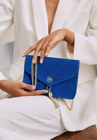 Borsa a tracolla blu in suede con catena dorata, chiusura a patta, dettaglio del logo e accentuazione con un pon pon. Forma rettangolare con una texture morbida.