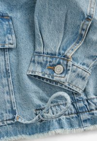 Veste en denim bleu clair avec fermeture à boutons, coutures visibles et ourlet effiloché. Le tissu présente une texture décolorée et un léger aspect usé.