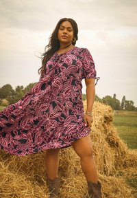 Eine Frau, die ein pinkes und schwarzes Blumenkleid sowie Stiefel trägt, steht selbstbewusst neben einem Heuhaufen in einem grasbewachsenen Feld unter einem bewölkten Himmel.
