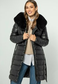 Zwarte pufferjas met een grote zwarte nepbontkraag, voorzien van een ritssluiting, glanzende textuur en gestikt ontwerp. Gekocht over een beige trui.