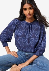 Donna seduta con le gambe incrociate, indossa una blusa blu a fantasia con maniche a sbuffo e jeans blu, guardando con sicurezza verso la fotocamera.