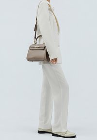 Braune Lederhandtasche mit strukturiertem Design, goldfarbenen Beschlägen, Tragegriff und abnehmbarem Schulterriemen. Getragen zu einem weißen Anzug.