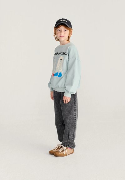 Bambino con i capelli rossi che indossa una felpa azzurro chiaro con disegni di fantasmi, cappellino nero, jeans grigio scuro e sneakers marroni, in piedi davanti a uno sfondo semplice.