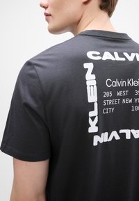 Μαύρο βαμβακερό μπλουζάκι με έντονο λευκό κείμενο στο πίσω μέρος, περιλαμβάνοντας "CALVIN KLEIN" και μια διεύθυνση σε μοντέρνο, γραφικό σχέδιο.