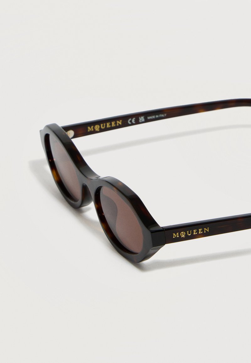 Schildpatt-ovale Sonnenbrille mit dunklen Gläsern und "McQueen" in Gold an den Bügeln eingraviert, vor einem schlichten weißen Hintergrund.