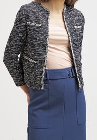 Marinblå texturerad jacka med ett vävt mönster, med metalltråd accenter, rund krage och två framfickor. Bärs över en beige topp.