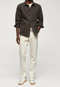 Man in een donkerbruin button-up shirt over een wit t-shirt, crème broeken en witte sneakers, staand tegen een effen achtergrond.
