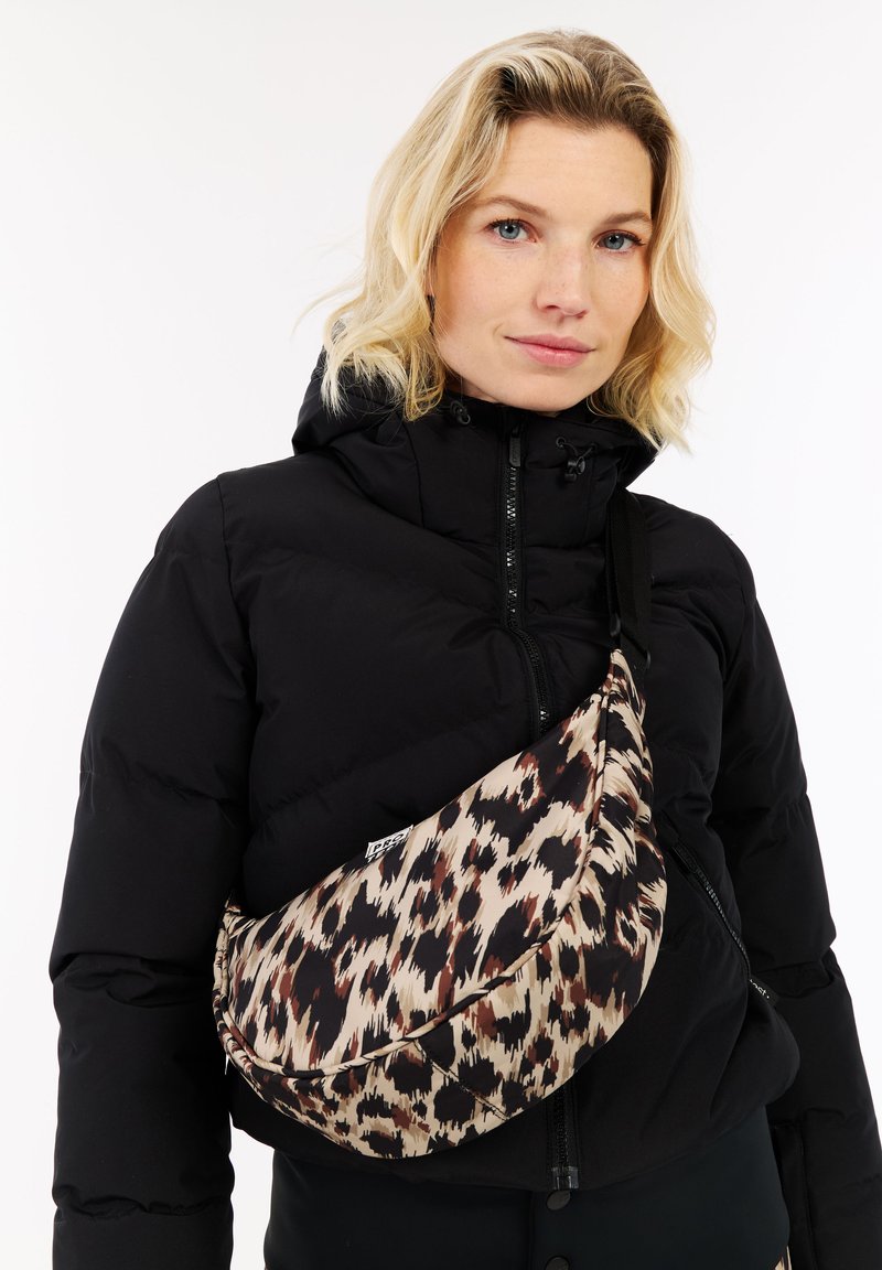 Blonde vrouw in een zwarte pufferjack met een crossbody tas van luipaardprint, kijkt recht vooruit met een neutrale uitdrukking.