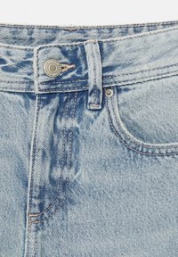 Gros plan de la ceinture d’un jean en denim bleu clair avec un bouton en métal et des coutures visibles autour de la poche et des passants de ceinture.