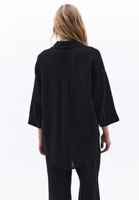 Czarna lniana koszula z kołnierzykiem, rękawami 3/4 i zaokrąglonym dołem. Posiada plisę z tyłu oraz mankiety na guziki. Prosty design.