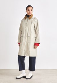 Light beige waterproof coat with a brown suede collar, two front pockets, and a zip closure, paired with blue jeans and white shoes.