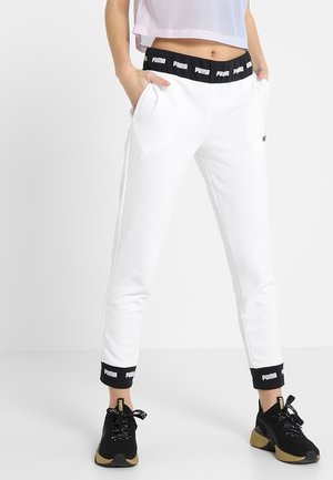 Pantalon de survêtement blanc Puma avec ceinture et poignets noirs à logo, porté avec des baskets noires Puma, modèle debout les mains dans les poches.