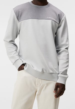 Man draagt een lichtgrijze en grijze J.Lindeberg colorblock trui met handen in de zakken van crèmekleurige broek.