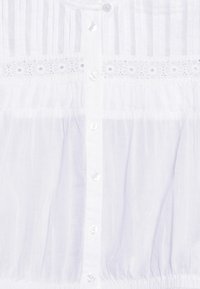 Blusa bianca realizzata in tessuto leggero, con strisce verticali, chiusura con bottoni e dettagli in pizzo sul petto e in vita.