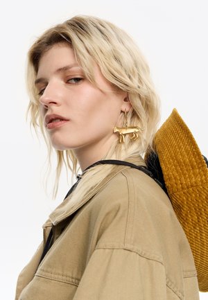 Chaqueta oversized beige con un cuello negro, que incluye un pendiente en forma de gato dorado y una mochila texturizada de color mostaza. Cabello liso, fondo neutro.